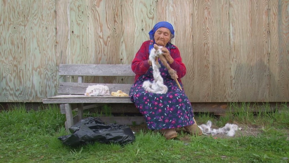 Photo : une dame âgée en robe mauve et gilet de laine rosée, assise sur un banc de parc, travaille un morceau de fourrure et de cuire. Derrière elle, un mur en bois contreplaqué.